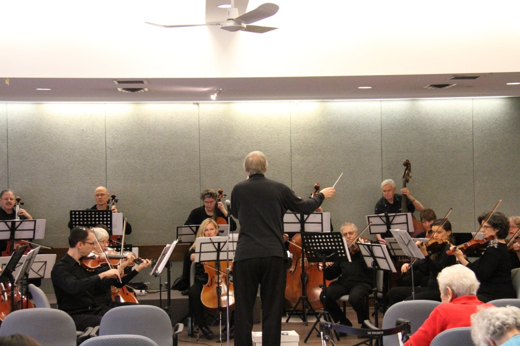 Conductor Matthew Poon leading Strings Attached Orchestra at a community concert at The Terraces of Baycrest, seen from the audience
