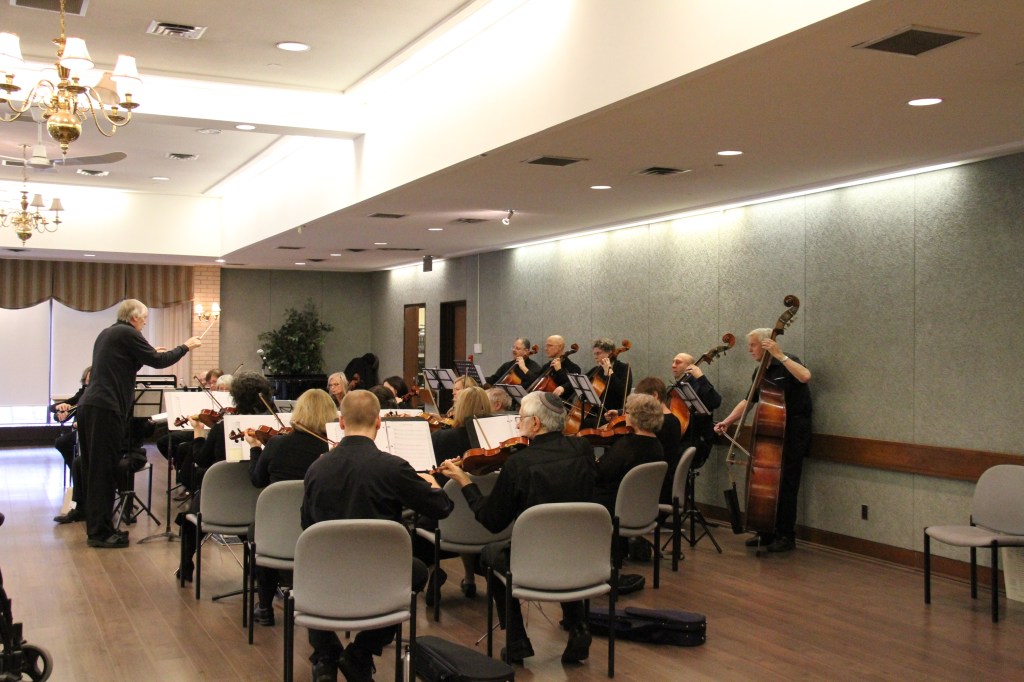 Wide view of the orchestra performing in a bright hall at The Terraces of Baycrest