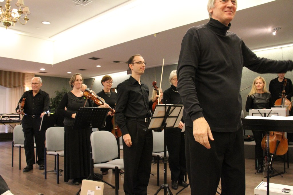 Conductor Matthew Poon addressing the audience while the musicians stand at the Baycrest community concert