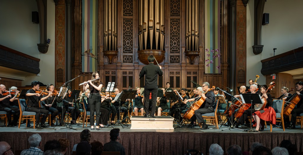 The full orchestra performing in front of the pipe organ at Trinity-St. Paul’s Centre, with the conductor on the podium