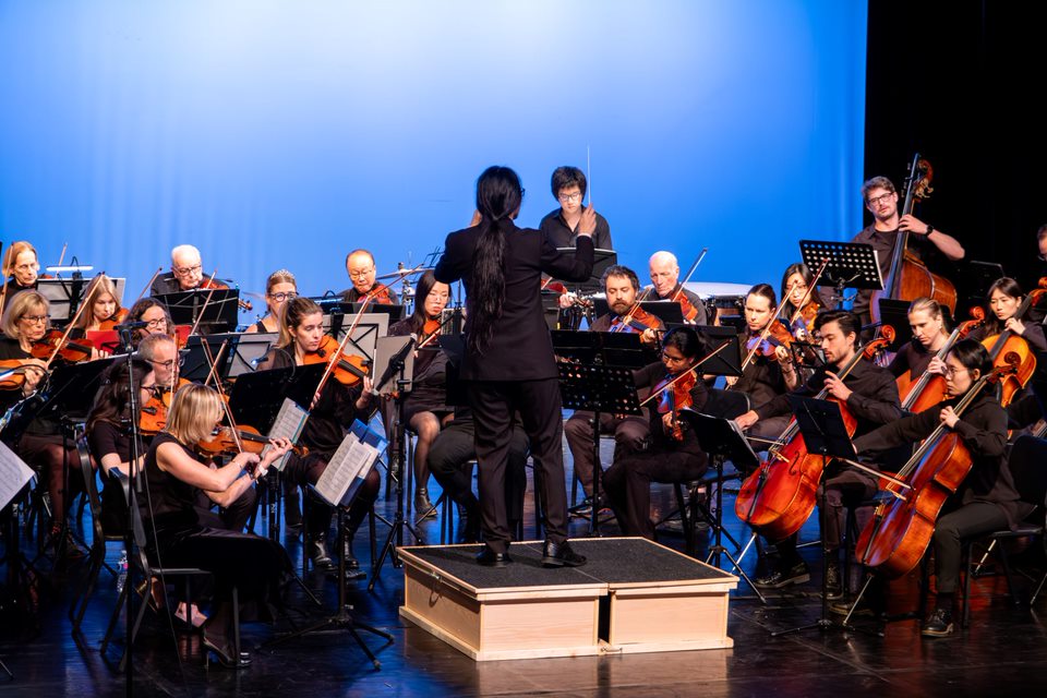 Jonathan Wong conducting the full Strings Attached Orchestra from the podium on a blue-lit stage