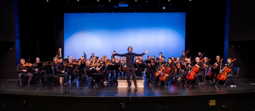 Strings Attached Orchestra on a blue-lit stage, with Music Director Jonathan Wong at the front, arms outstretched toward the audience