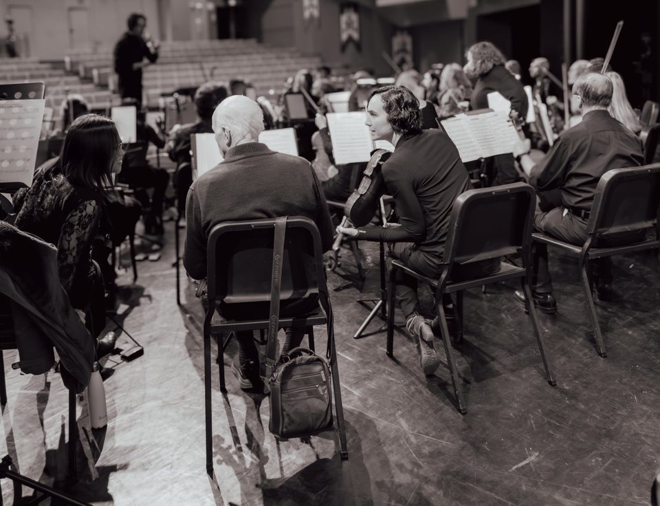 Black-and-white photo of the orchestra on stage, seen from behind the players