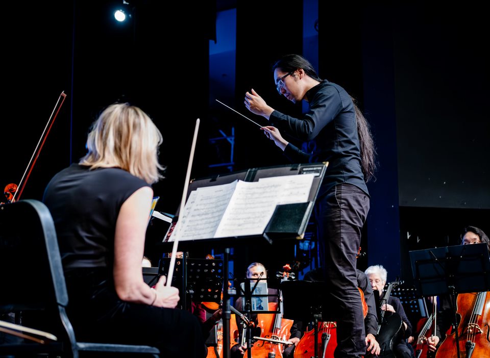Jonathan Wong conducting, seen past a string player and her music stand