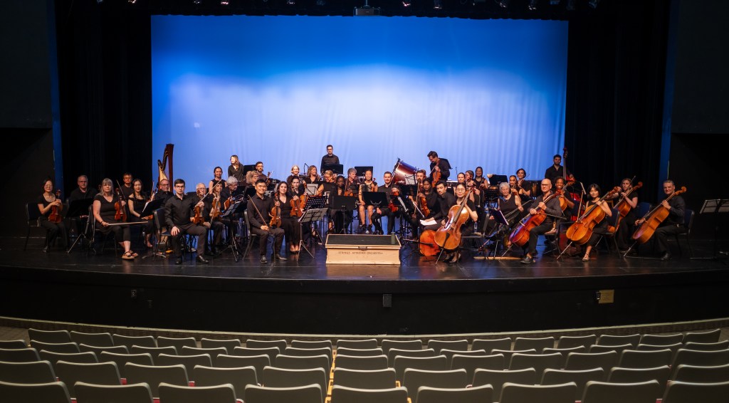 The full orchestra on stage before a blue backdrop, seen from the empty hall
