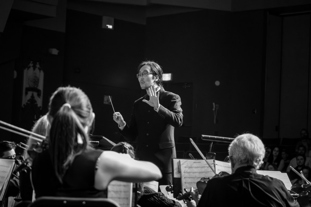Black-and-white photo of Jonathan Wong conducting, seen past the strings
