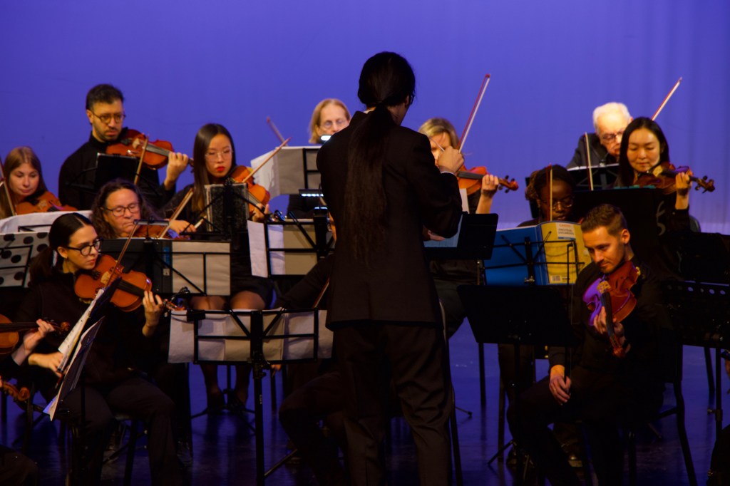 Jonathan Wong, seen from behind, conducting the violins on a blue-lit stage