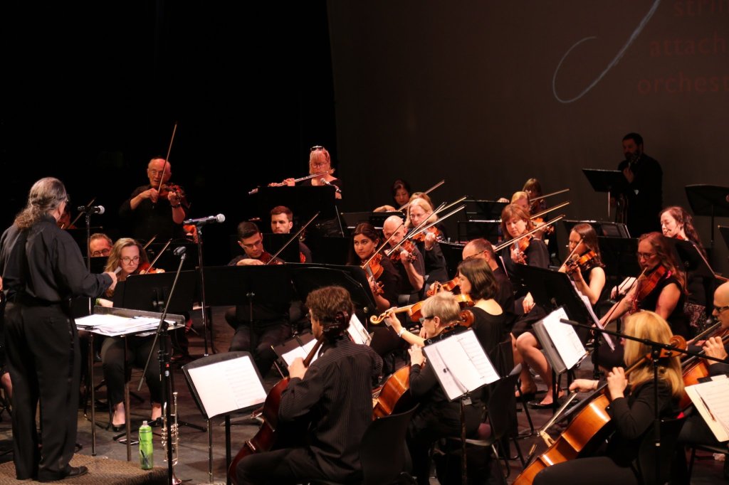 Strings Attached Orchestra performing on a dark stage at the Isabel Bader Theatre, with the conductor at the left