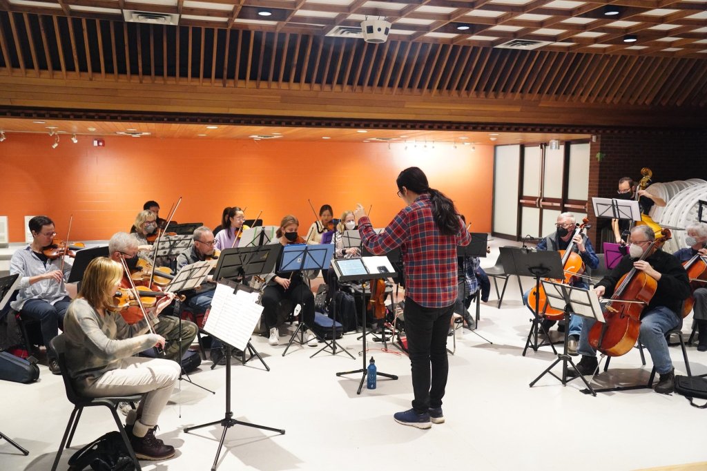 The orchestra rehearsing in a hall with an orange wall and a wooden ceiling