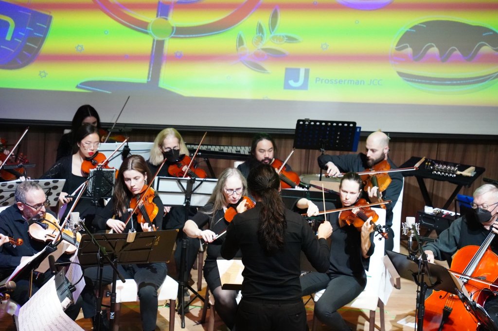 The orchestra on stage at the Leah Posluns Theatre beneath a projected Hanukkah menorah